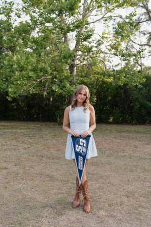 Young woman in white dress and cowboy boots holding a blue college pennant flag outdoors on a sunny day.