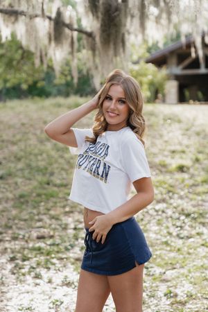 Young woman in white crop tee and navy shorts posing outdoors under Spanish moss-draped trees.