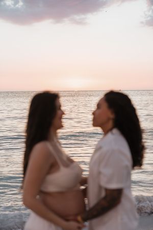 Two women face each other holding a pregnant belly at sunset by the ocean in a romantic maternity photo.
