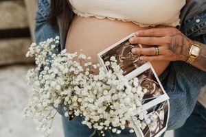 Pregnant woman holding baby's breath flowers and ultrasound photos against her bare baby bump, with a tattooed hand visible.