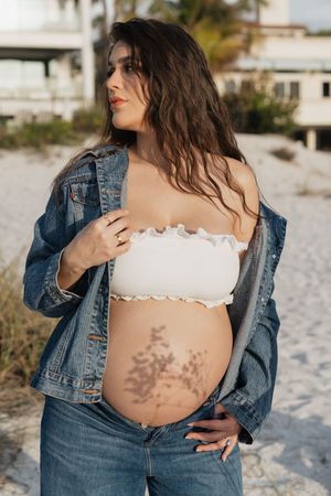 Pregnant woman in white bandeau top, denim jacket, and jeans poses on sandy beach, showing baby bump with floral shadow.