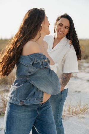 Two women smiling and laughing together on a sandy beach, one in a denim jacket and the other in a white shirt.