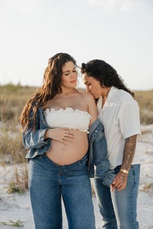 Pregnant woman in white tube top and jeans holds her belly as partner leans in tenderly on a sandy beach.