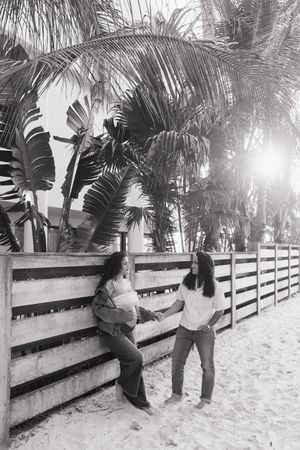 Black and white photo of two women conversing by a wooden fence on a sandy beach surrounded by tropical palm trees.