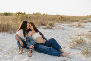 Two women share a tender moment on sandy beach dunes, one pregnant, both wearing jeans, surrounded by golden grass.