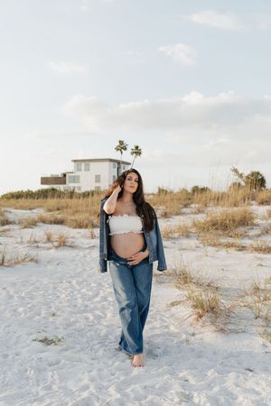 Pregnant woman in white crop top and jeans poses barefoot on sandy beach with palm trees and white building behind her.