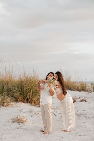 Couple in white outfits holding wildflowers on sandy beach dunes with sea grass and cloudy sky backdrop.
