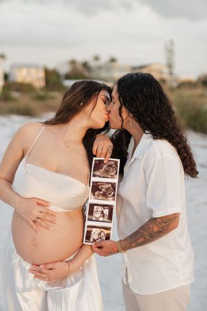 Two women kissing on a beach, one pregnant holding her belly, both holding ultrasound photos outdoors.