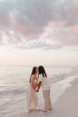 Two women share a tender moment on a sandy beach at sunset, one visibly pregnant, both dressed in white.