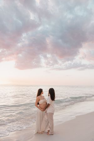 Two women stand together on a serene beach at sunset, one pregnant, gazing at the pastel-colored sky over the ocean.