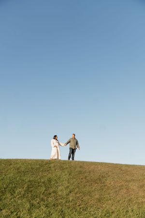 A couple walks together on a grassy hilltop against a bright blue sky, creating a silhouetted romantic scene.