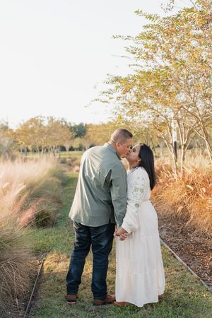 A couple in romantic poses outdoors during golden hour with tall grass and autumn trees in the background.