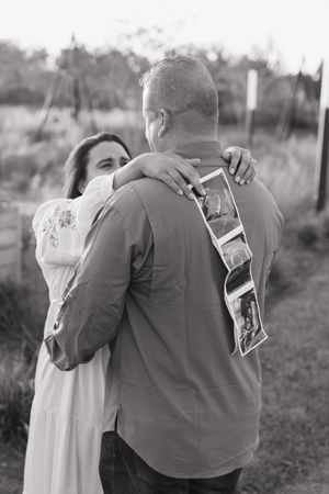 A couple embraces while holding photographs during an outdoor photoshoot in black and white.