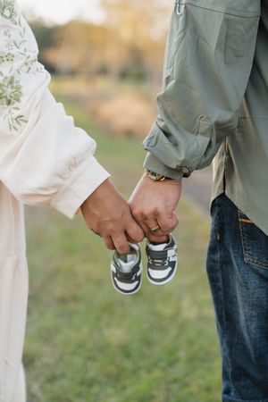 Couple holding small baby shoes together in an outdoor setting with grass in the background.