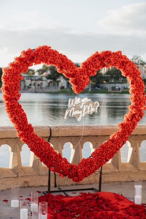 Heart-shaped red rose petal arch decorates a waterfront proposal setup with romantic candles and decor.