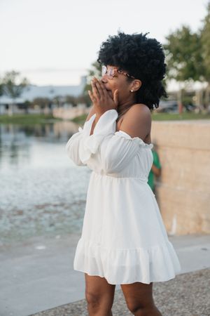 Person in a flowy white off-shoulder dress laughs joyfully by a waterfront on a sunny day.