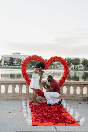 A romantic proposal setup with a heart-shaped arch made of red roses by a lakeside balustrade.