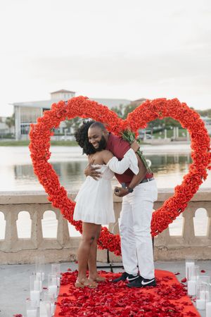 A couple embraces under a large red floral heart arch with rose petals and candles on a balcony overlooking water.