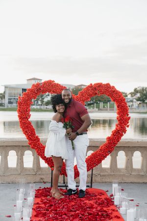 A couple poses together beneath a red rose heart-shaped arch with rose petals and candles decorating the ground.