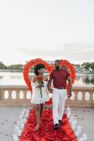 A romantic proposal setup on a waterfront balcony with a heart made of red rose petals and white candles along the path.