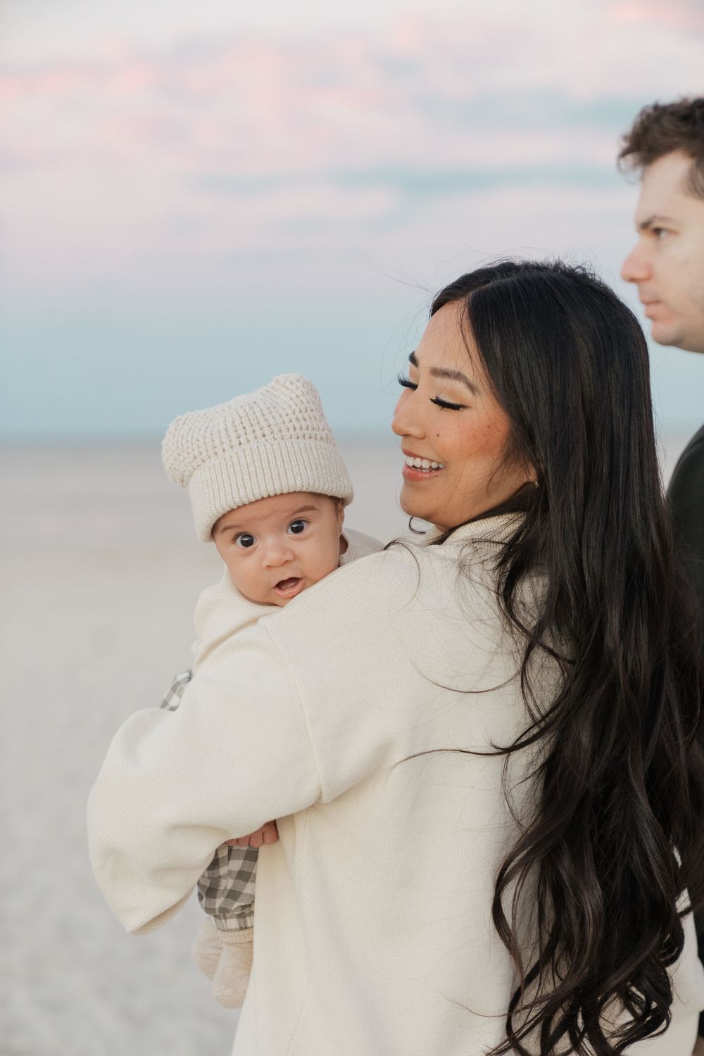 A mother and baby in matching cream sweaters share tender moments together on a beach at sunset.