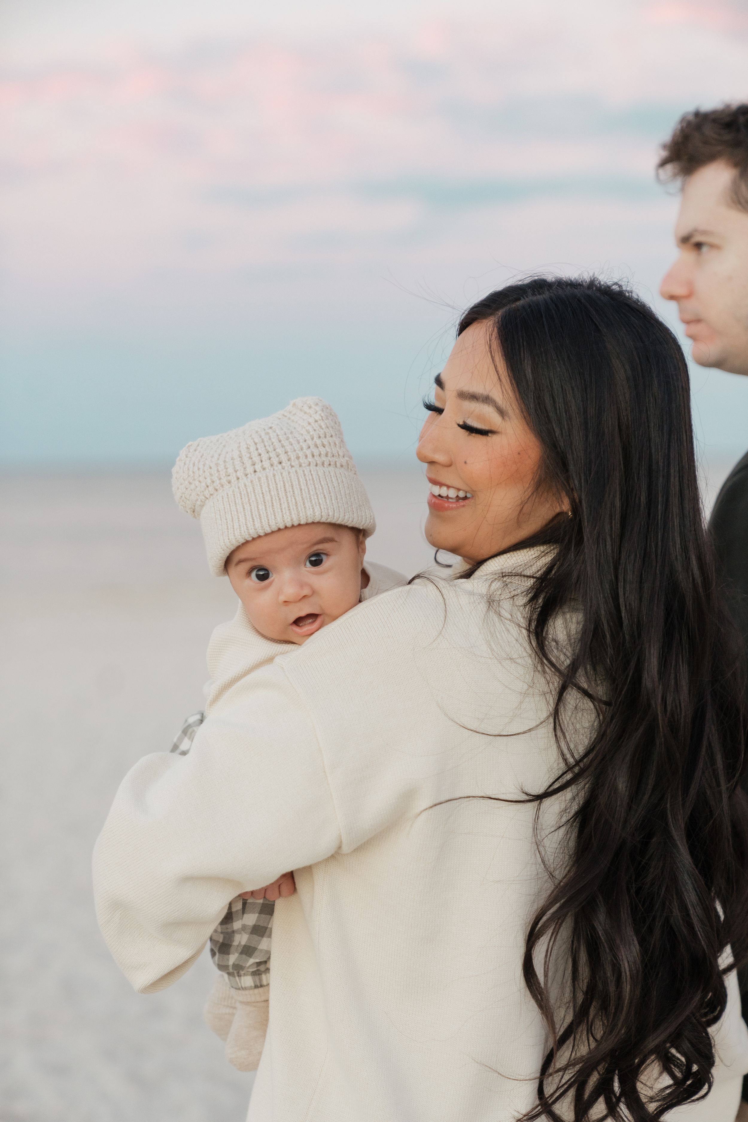 A mother and baby in matching cream sweaters share tender moments together on a beach at sunset.