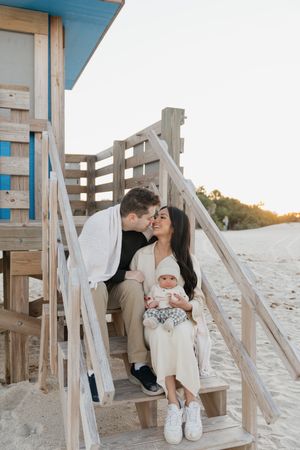 A romantic couple shares a tender moment on wooden beach stairs at sunset while wearing white outfits.