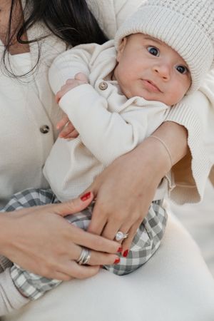 Baby wearing white knit hat and sweater lies on soft white bedding while being gently held by adult hands.
