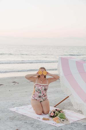 A beachside picnic scene with a pink striped blanket, flowers, and watermelon on a sandy shore at sunset.