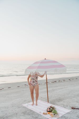 Pink striped beach umbrella and picnic blanket set up for a romantic sunset date on a sandy beach.