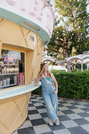 Person in denim overalls poses against a checkered floor and ice cream shop backdrop in a sunny outdoor setting.