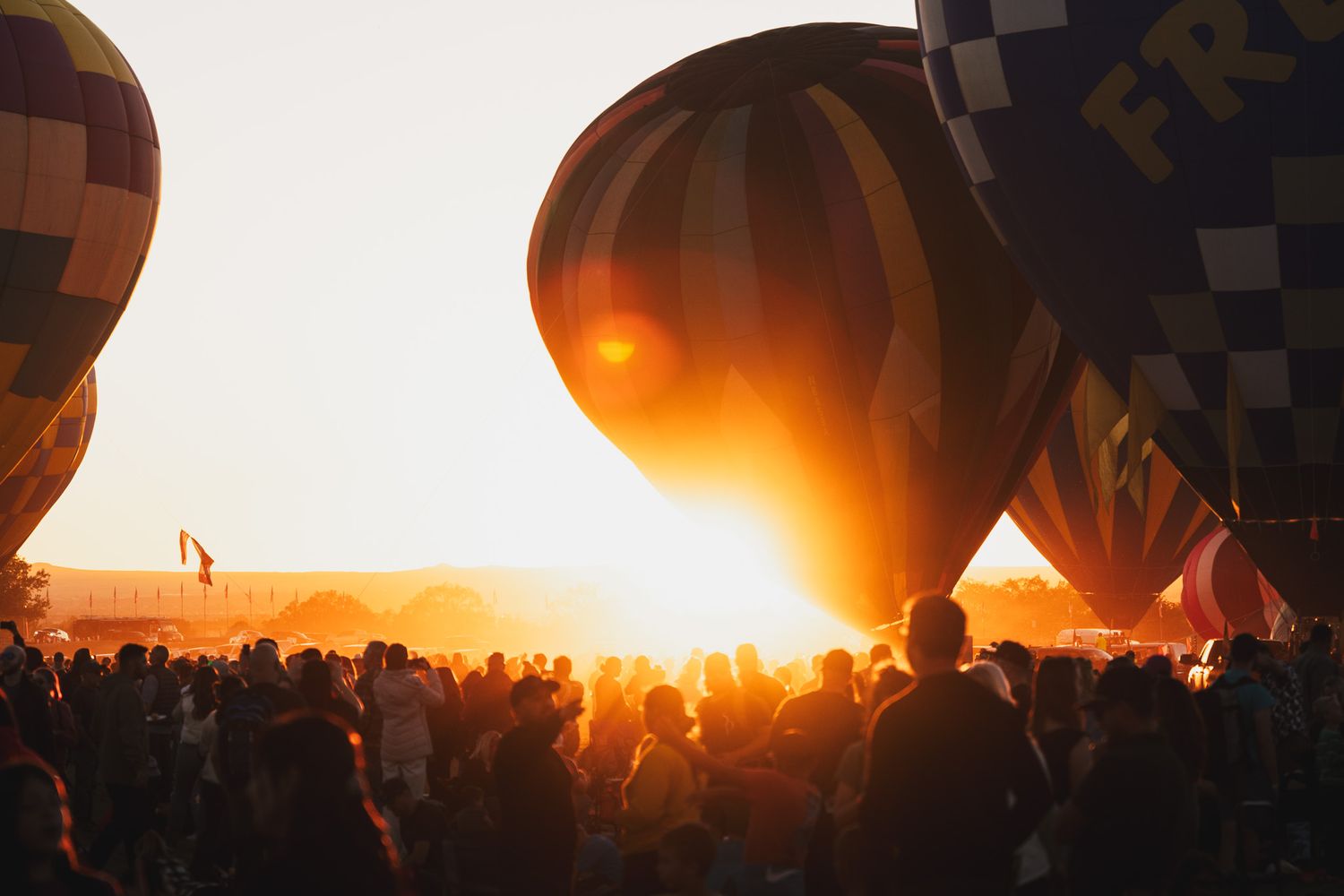 Hot air balloons silhouetted against bright sunset with crowd gathered below.