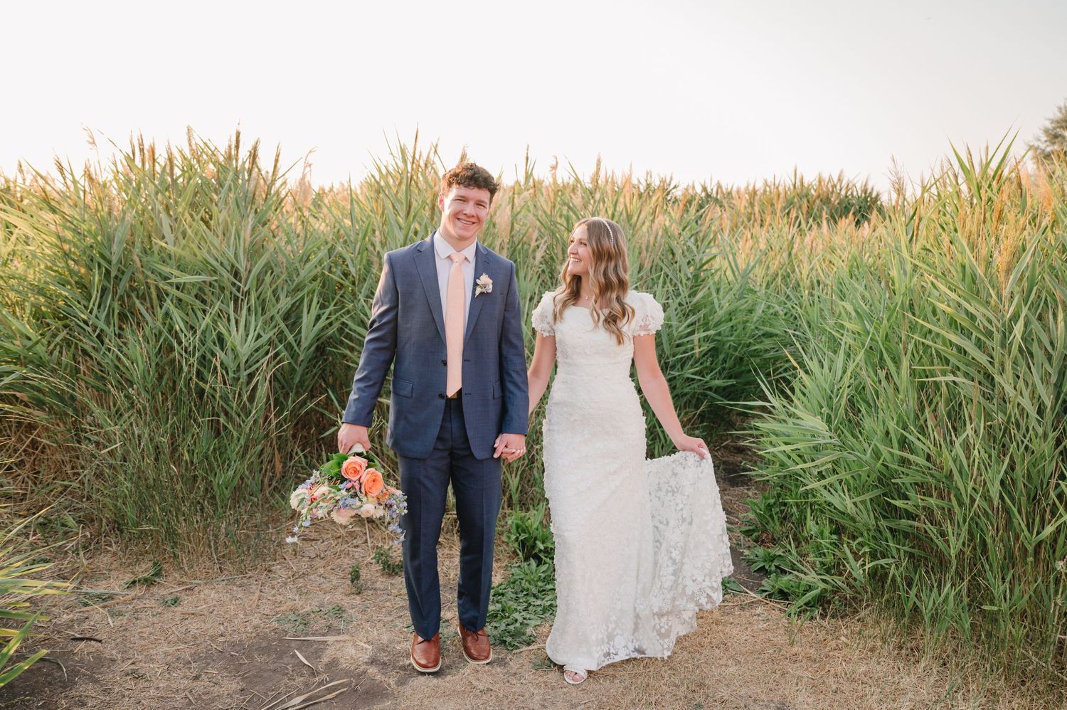 Newlyweds holding hands while walking along path through tall grass.