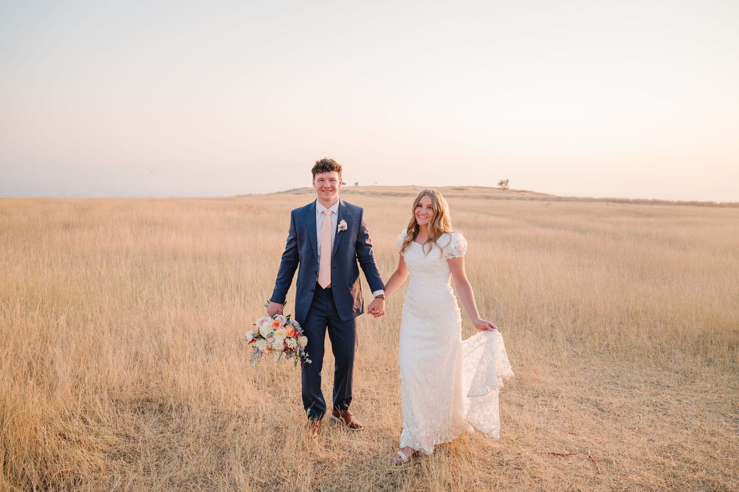 Newlyweds walking together through golden grass field during sunset.