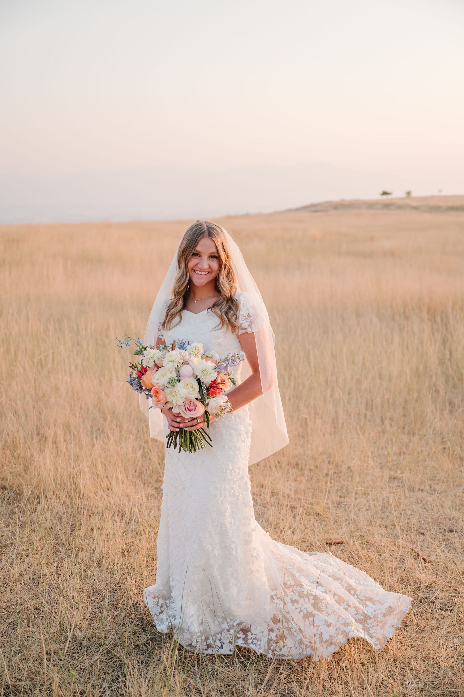 Bride in lace wedding dress holds colorful bouquet while standing in golden field at sunset.
