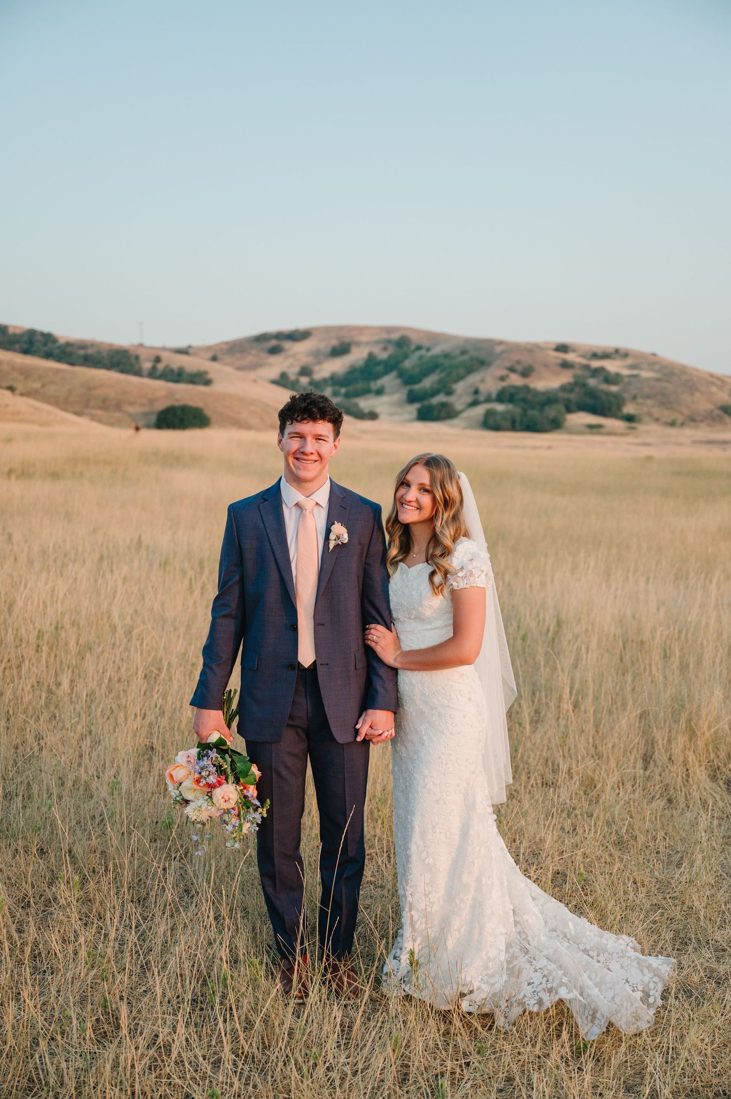 Newlyweds pose together in golden field with rolling hills during sunset.