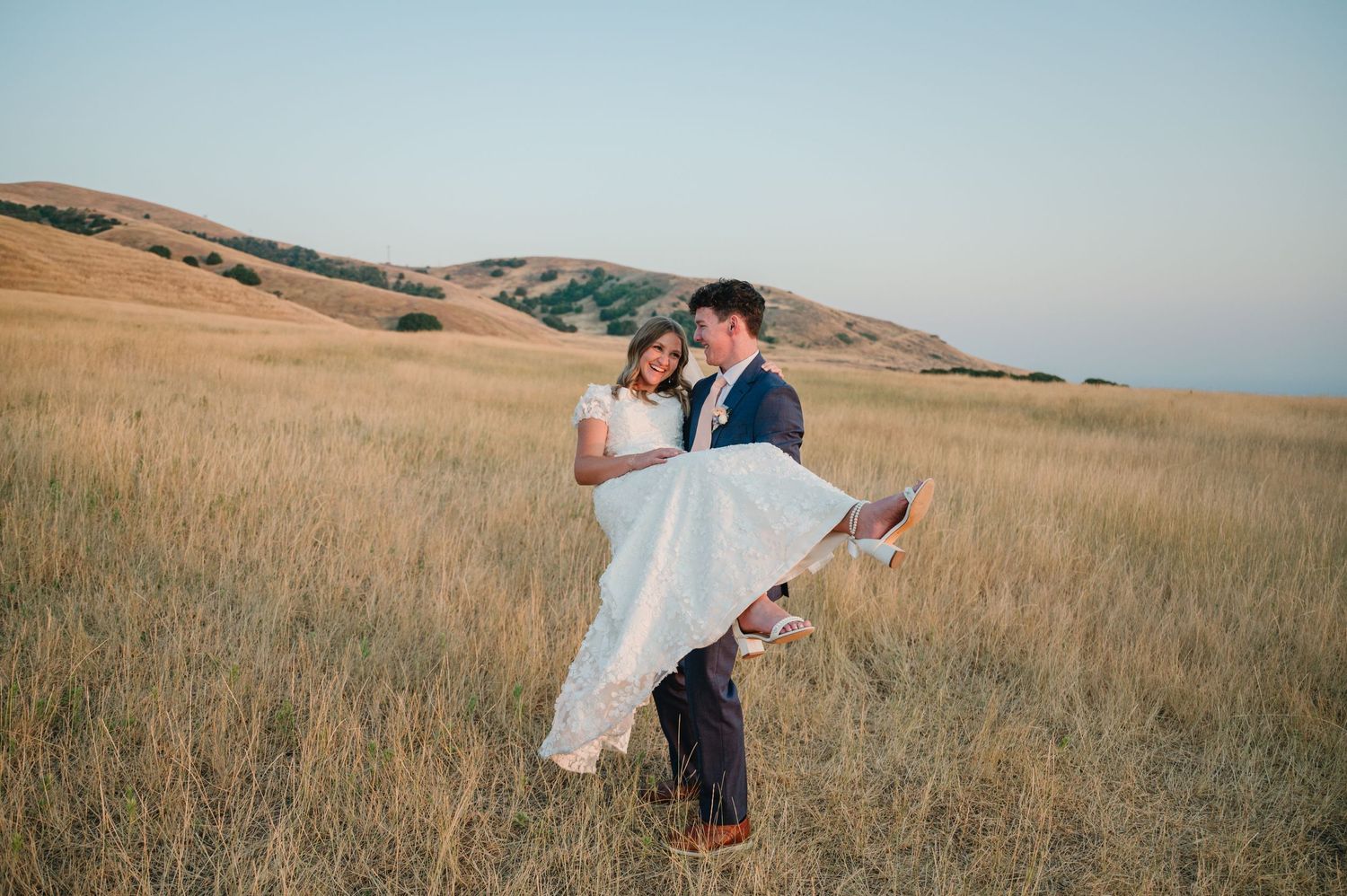 Romantic couple embraces in grassy field during sunset with rolling hills in background.