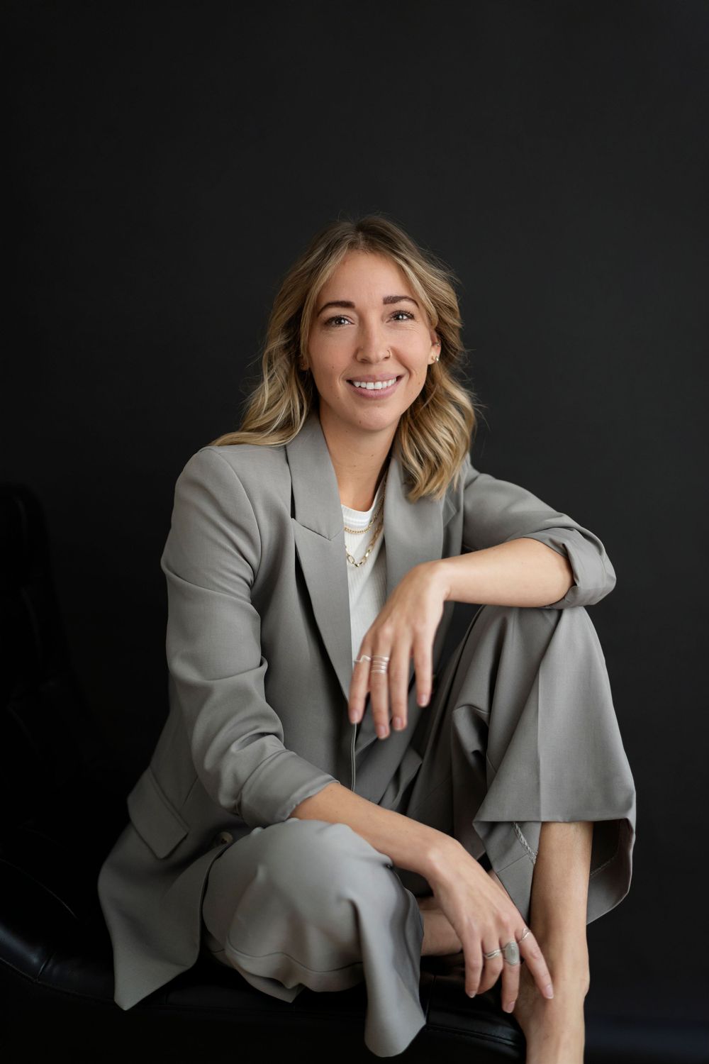 Professional portrait of woman in grey suit seated casually against dark background.