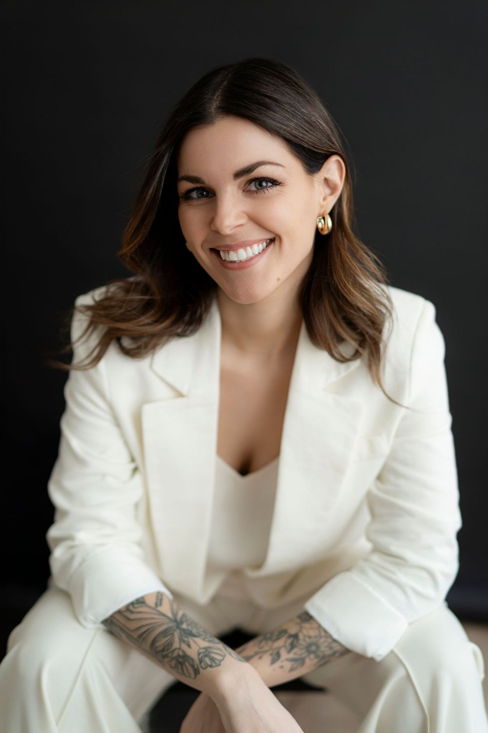 Professional portrait of woman in white blazer with tattoos against dark background.