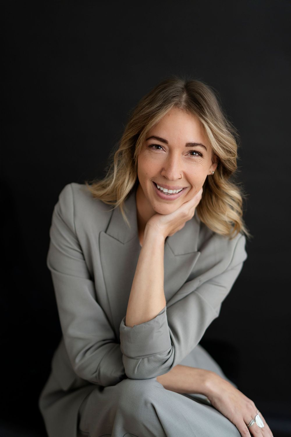 Professional headshot of a person in a gray blazer smiling warmly against a dark background.