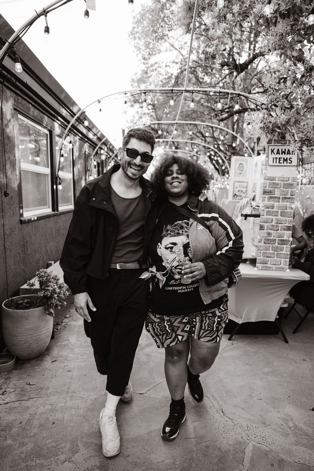 A black and white photo of two people posing together outdoors at a casual venue with string lights overhead.