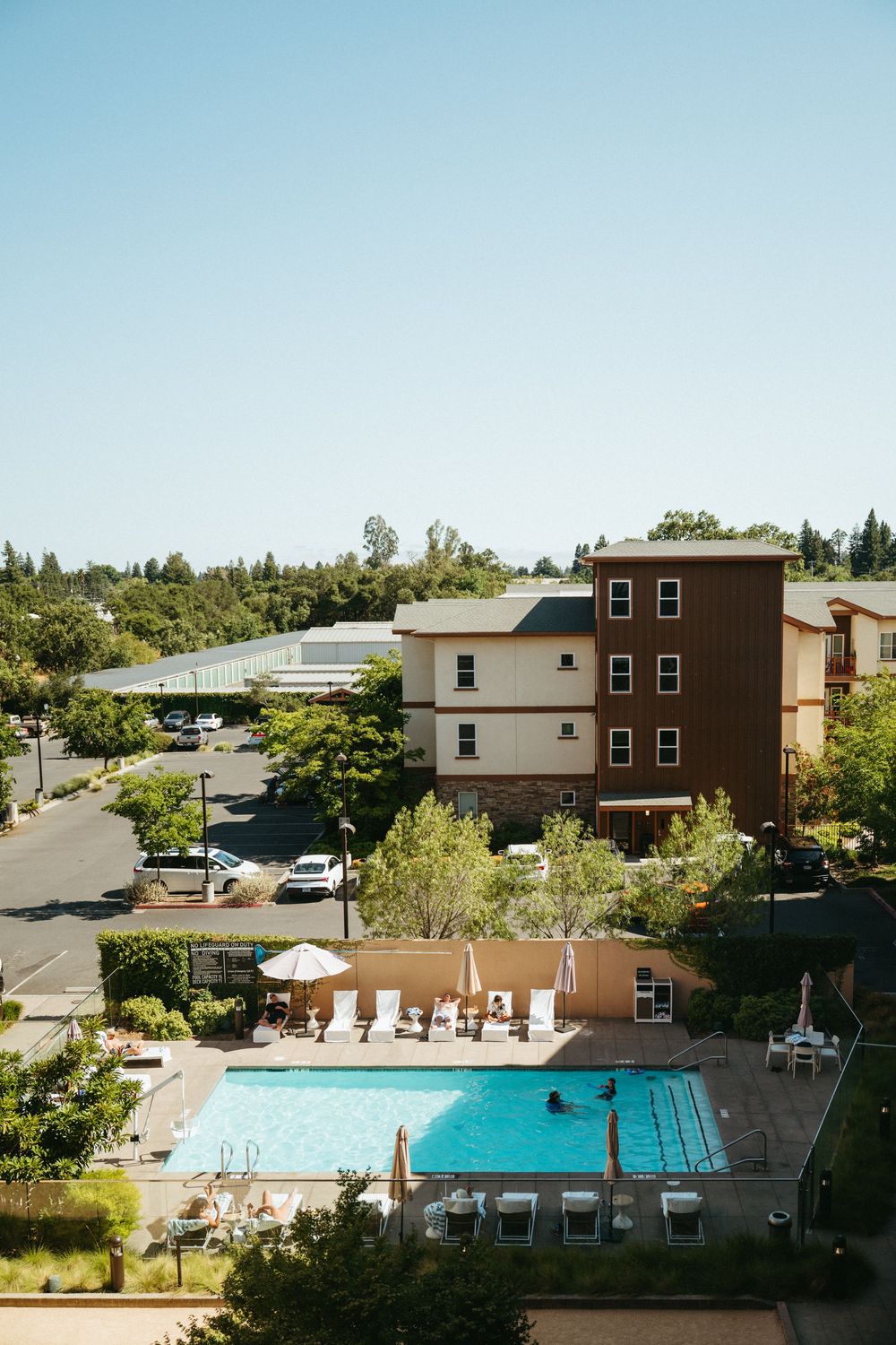 Aerial view of an apartment complex with outdoor swimming pool, lounge chairs, and surrounding buildings on a sunny day.