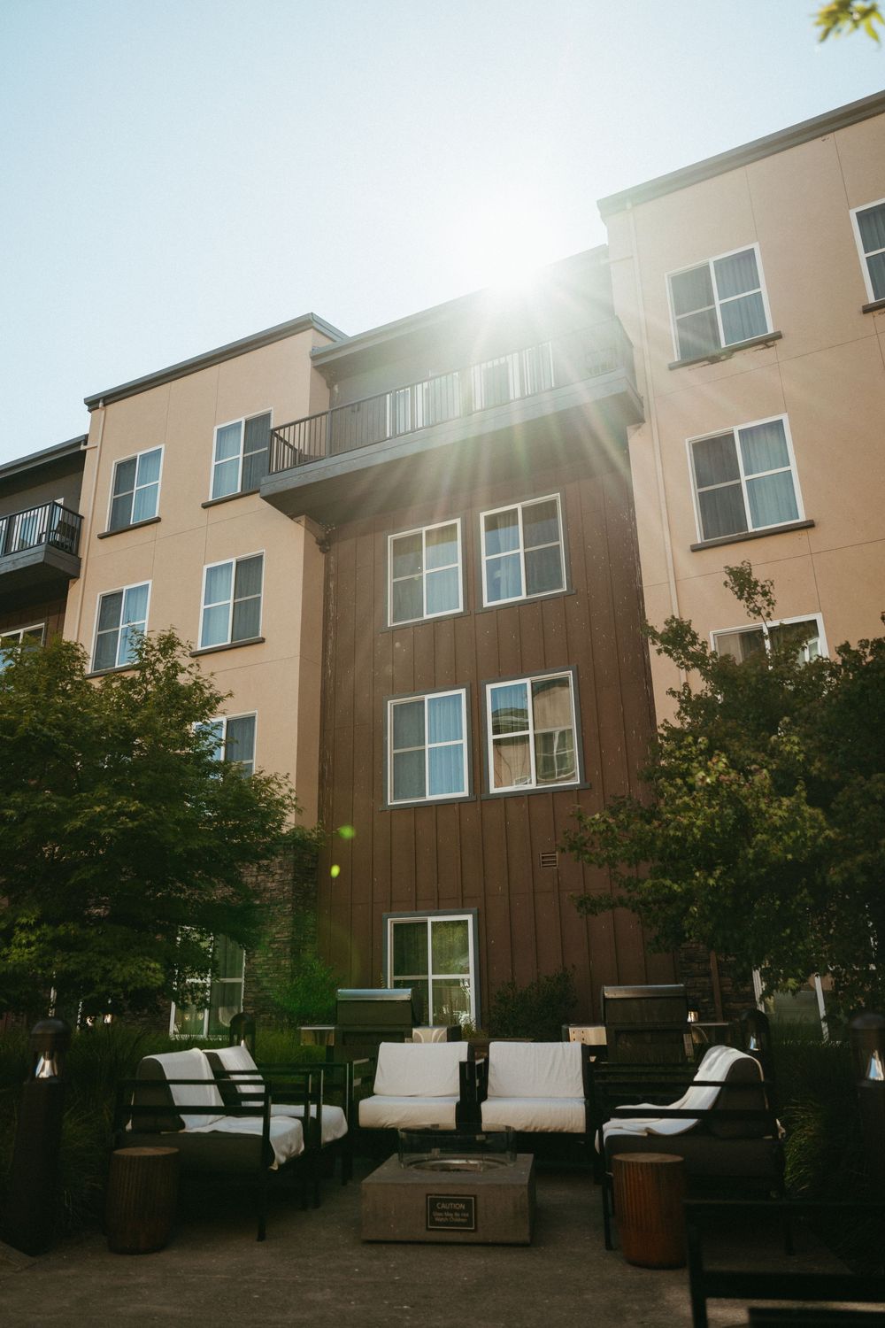 Modern beige apartment building exterior with large windows and trees in sunlight against blue sky.