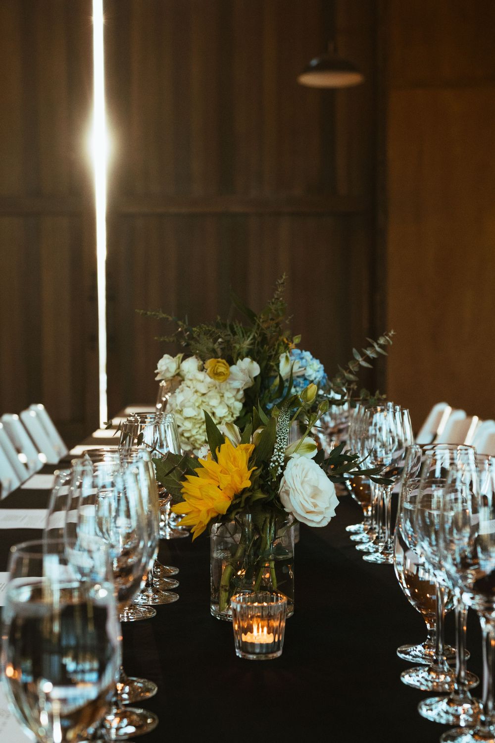 Elegant wedding reception table setting with wine glasses and sunflower floral centerpieces in a dark, moody atmosphere.