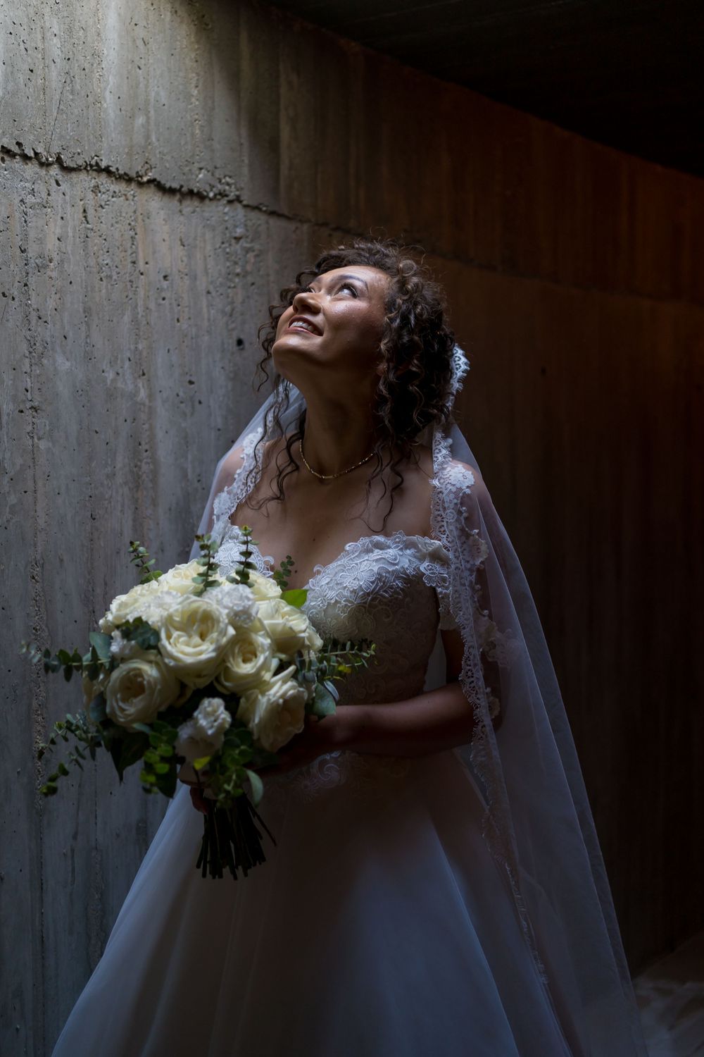 A bride in a lace wedding gown and veil holds a white rose bouquet while looking up in dramatic lighting.