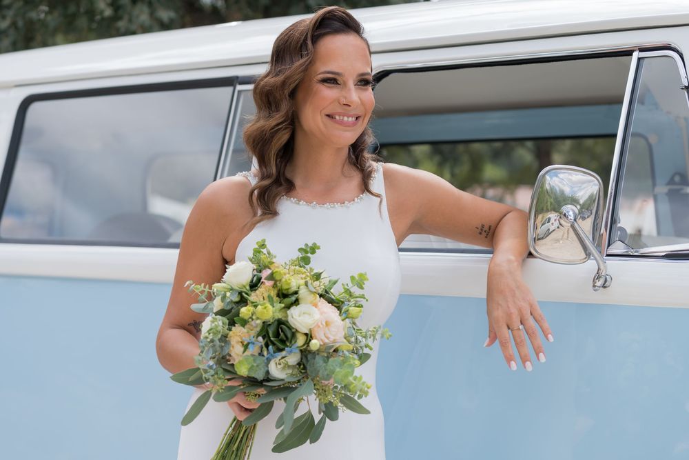 Bride in white dress holding floral bouquet leans against light blue vintage Volkswagen van on sunny day.