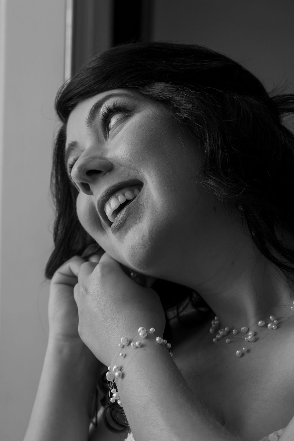 Black and white portrait of a joyful woman laughing with dark hair wearing delicate floral jewelry near a window.