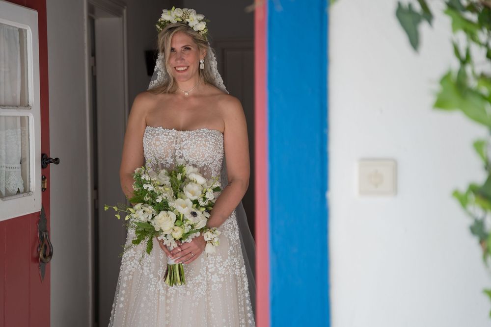 A bride stands in a doorway wearing a classic white strapless gown, flower crown and holding a white floral bouquet.