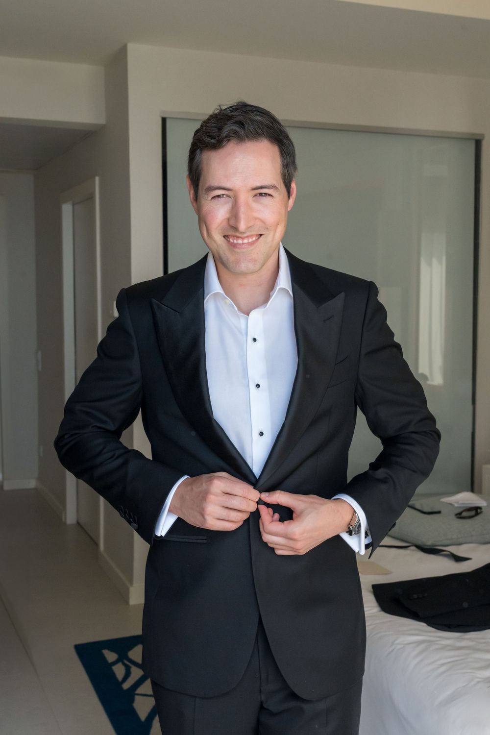 Professional wearing a classic black suit and white dress shirt buttons up while getting ready in a modern room setting.