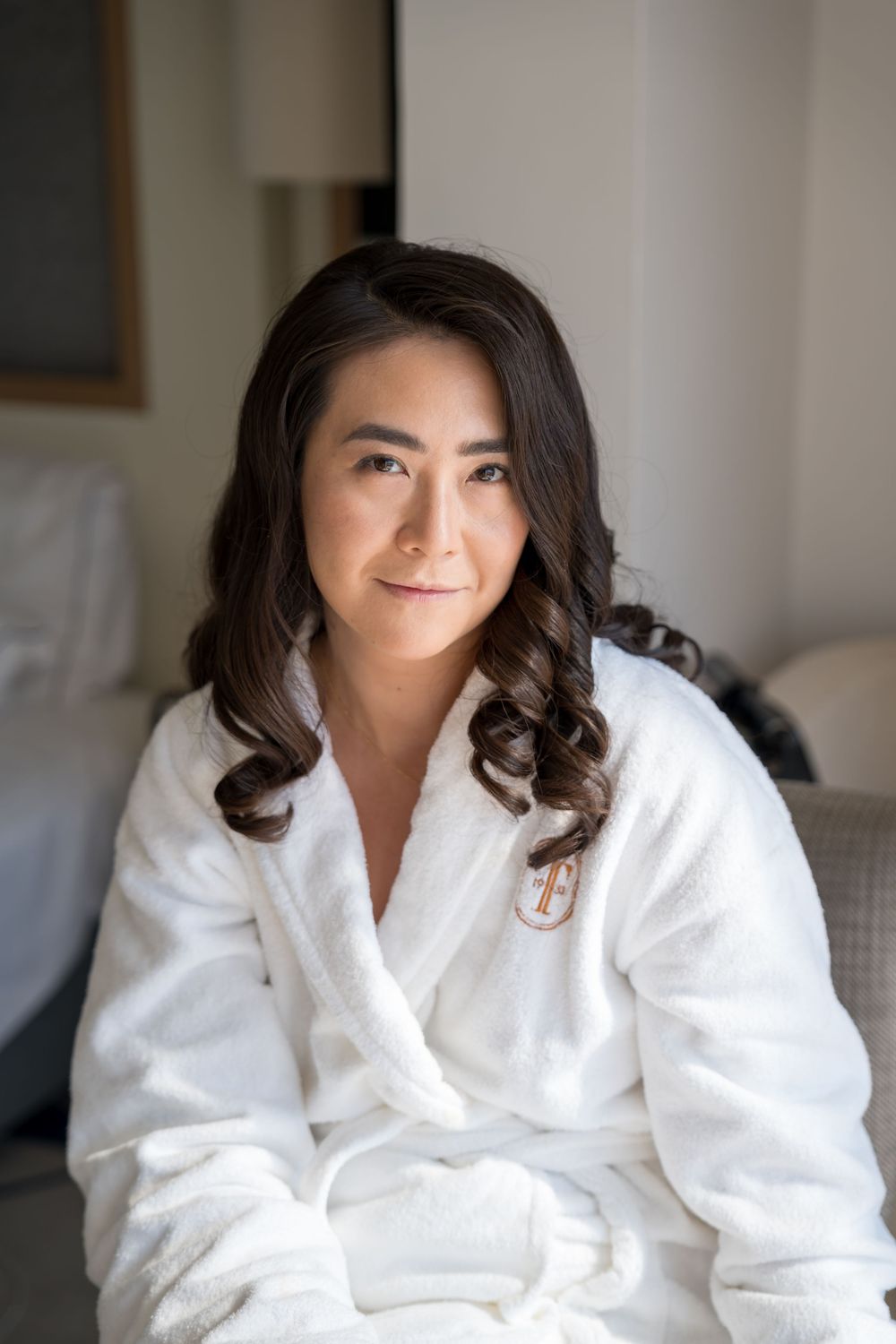Person in a white plush bathrobe with long dark wavy hair relaxing in a serene indoor setting.